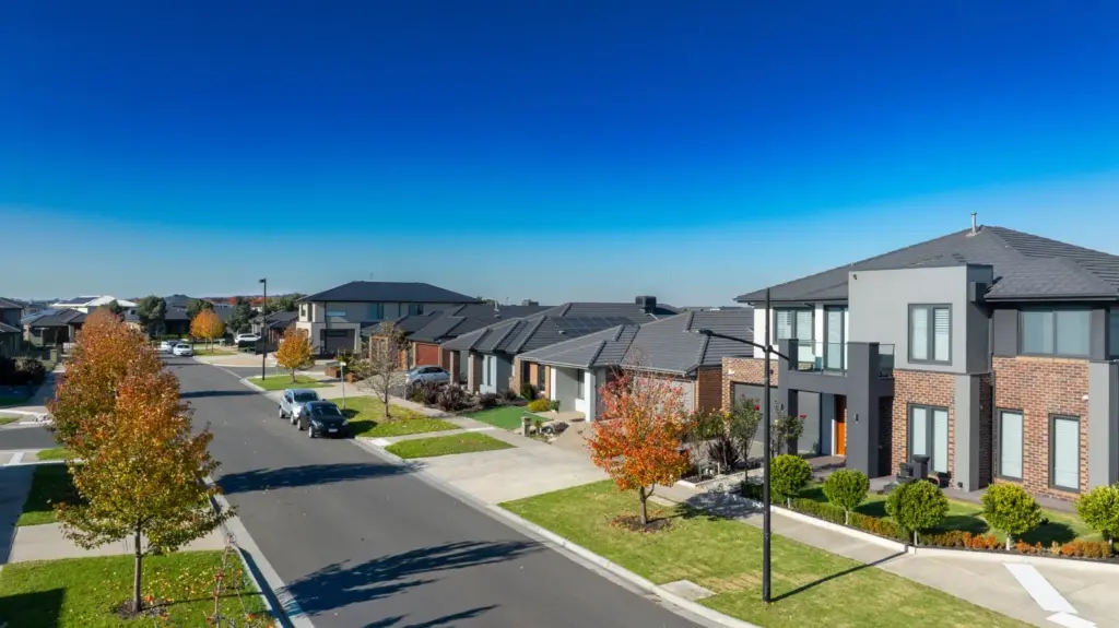 A suburban street with modern houses, neatly trimmed lawns, and a few cars parked along the road under a clear blue sky. Some trees display autumn foliage.