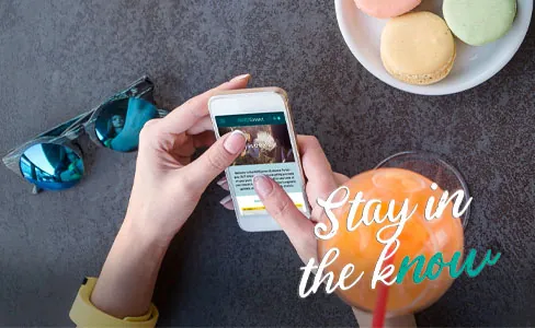 A person holds a smartphone and scrolls through an article at a table with sunglasses, colorful macarons on a plate, and an orange drink. Text on the image reads “Stay in the know.”.