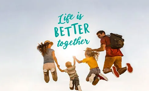 A family of four jumps in the air, holding hands, with the text “Life is better together” written above them against a clear blue sky.