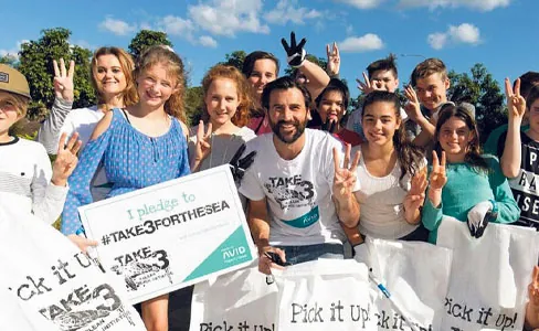 A diverse group of people, including children and adults, smile and hold up three fingers while participating in a #Take3ForTheSea cleanup event, holding bags and a pledge sign under a sunny sky.