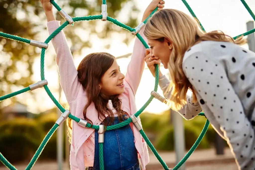 A young girl smiles while hanging from a rope climbing structure at a playground as a woman, possibly her mother, leans in close, smiling back at her. The background is outdoors with soft sunlight.
