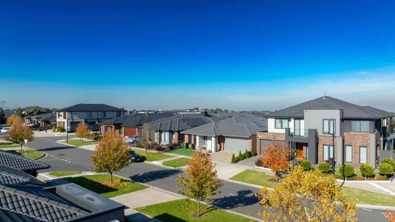 A suburban neighborhood with modern two-story homes, neatly trimmed lawns, and tree-lined streets under a clear bright blue sky. Some trees display autumn foliage with orange and yellow leaves.