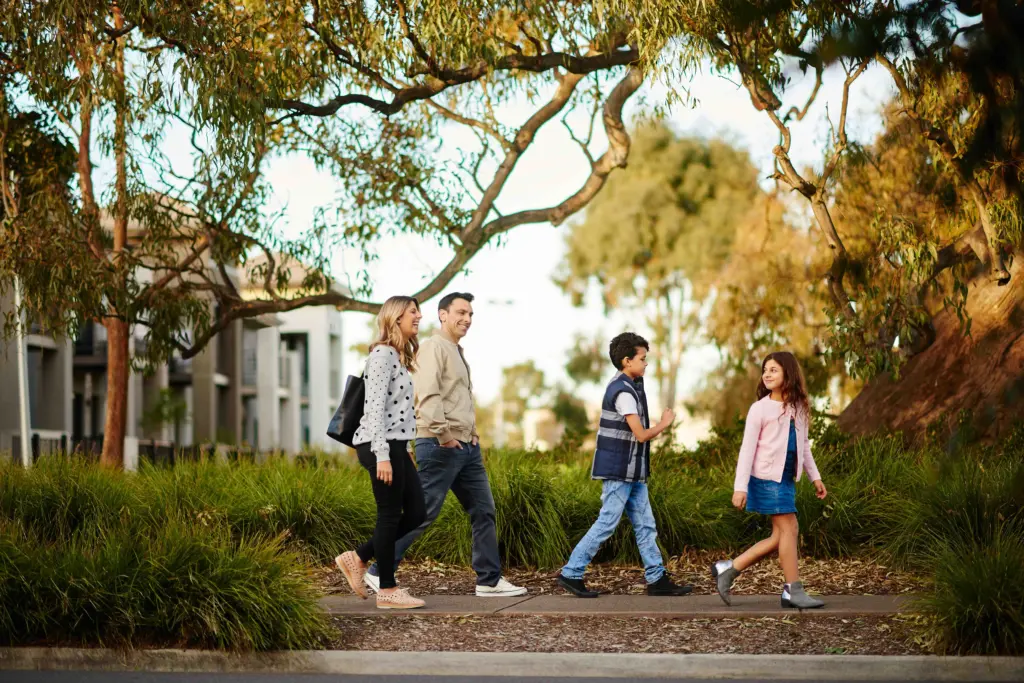 A family of four, including two adults and two children, walks together along a path surrounded by greenery and trees, with modern buildings visible in the background.