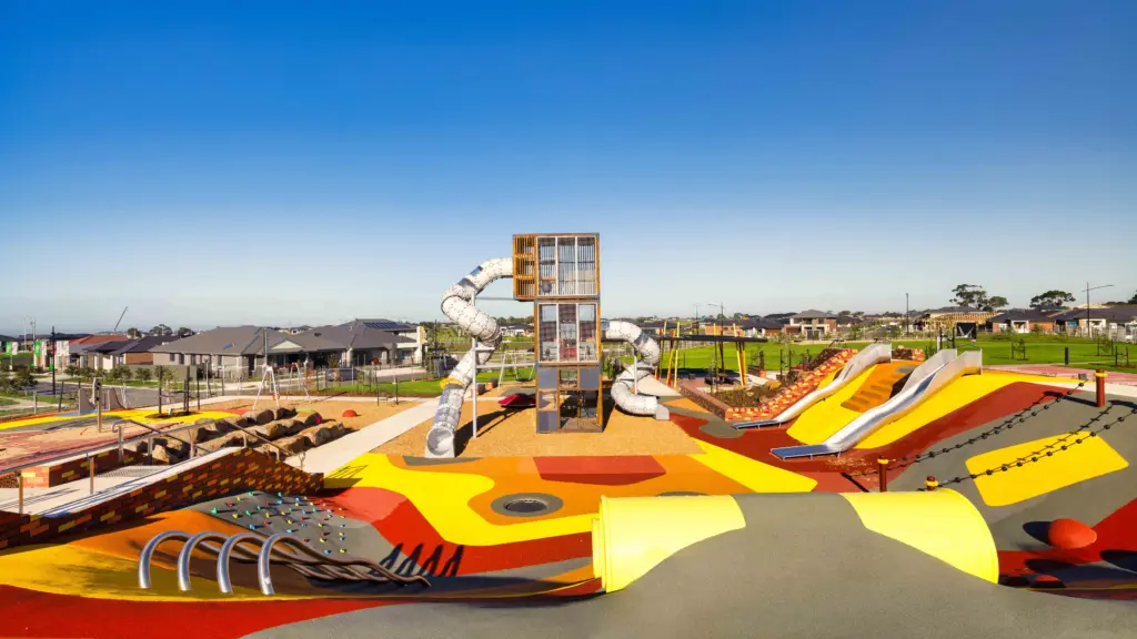 A colorful modern playground features slides, climbing structures, tunnels, and soft rubber flooring. Houses and a clear blue sky are visible in the background.