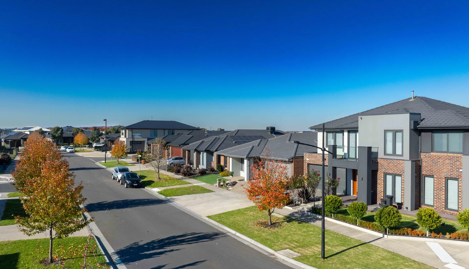 A suburban street lined with modern two-story houses, green lawns, and young trees with autumn leaves under a clear blue sky. A few cars are parked along the curb.